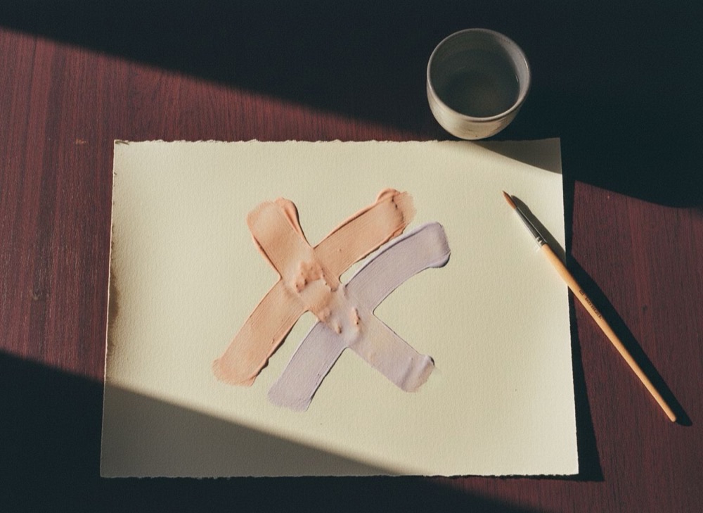 Two brush strokes, one peach and one lilac, crossing on a sheet of paper